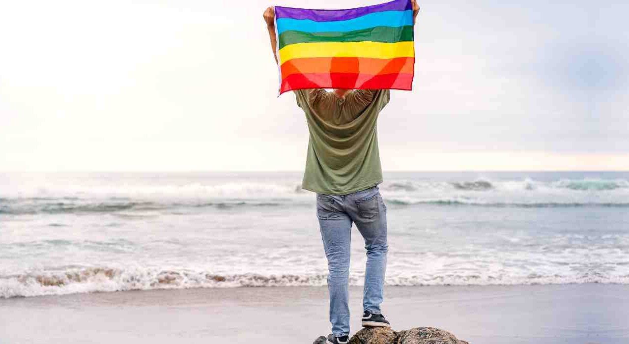 Youth Stands On Beach Holding Up LGBTQ+ Flag Which Depicts Wave Counselling And Wellness LGBTQ+ Affirming Therapy Services In St John's Newfoundland And Labrador