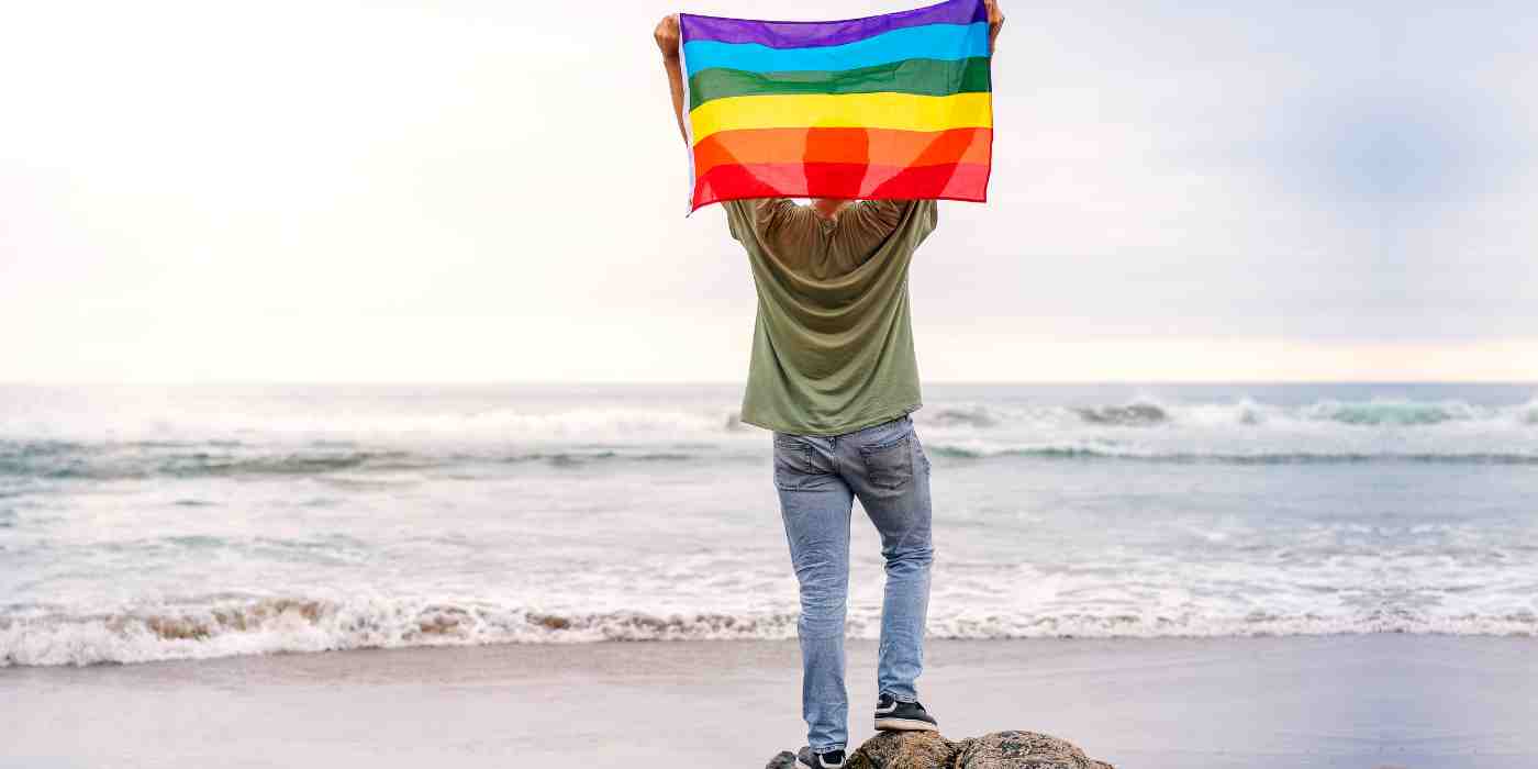 Youth stands on beach holding up lgbtq+ flag which depicts wave counselling and wellness lgbtq+ affirming therapy services in st john's newfoundland and labrador